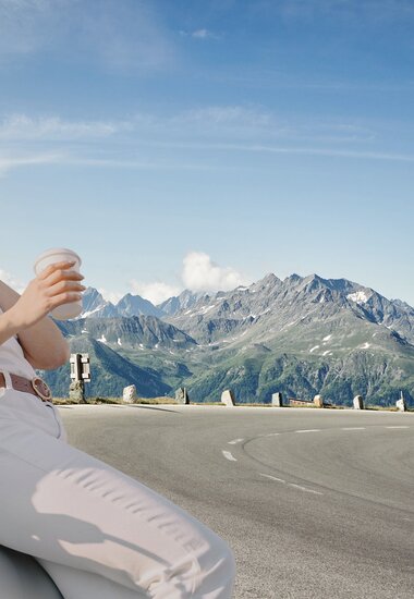 Frau sitzt am Auto mit Kaffee in der Hand und lächelt, Glocknerstraße im Hintergrund | © grossglockner.at